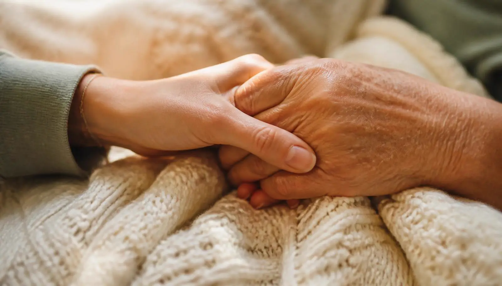 A younger hand gently holding an older hand on a cream blanket.